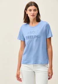 Camiseta de algodón color azul claro con mangas cortas, escote redondo y texto impreso "Todo se trata de la SENSACIÓN" en diferentes tamaños y estilos.