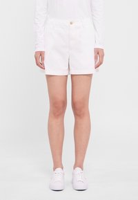 Shorts en coton blanc à coupe droite, avec poches avant et fermeture par un seul bouton métallique. Portés avec des baskets blanches.