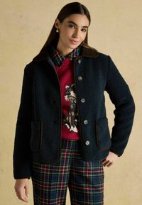 Giacca in pile blu navy con rifiniture marroni, chiusura a bottoni e due tasche frontali, indossata sopra un maglione rosso con un design di cane e pantaloni a quadri.