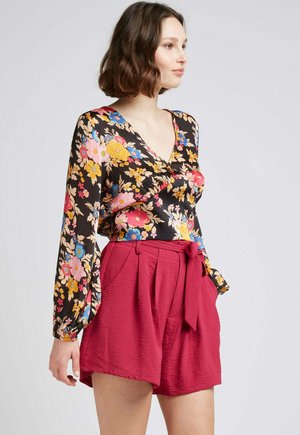 Blouse fleurie noire avec des fleurs vibrantes roses, jaunes et bleues ; décolleté en V et manches longues bouffantes. Associée à un short maroon taille haute.