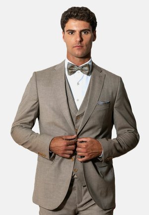 Man die een beige colbert dichtknoopt, met een bijpassend vest, wit overhemd en een patroonse vlinderstrik met pochet, neutrale achtergrond.