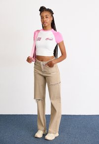 Top corto blanco con mangas rosas, que presenta logotipos; jeans de pierna ancha beige con roturas; zapatillas crema; modelo sostiene un bolso rosa, de pie sobre una alfombra azul.