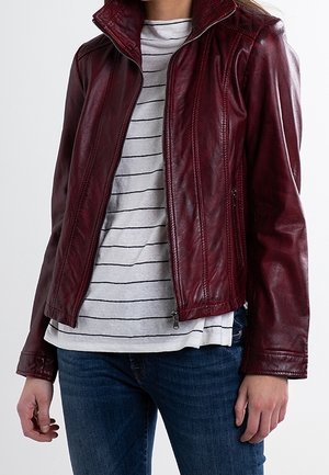 Personne portant une veste en cuir bordeaux sur une chemise blanche à rayures noires avec un jean bleu, debout, les bras détendus le long du corps.