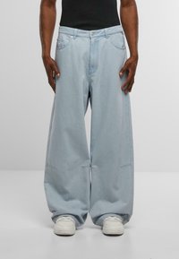 Pantalones de mezclilla de gran tamaño en azul claro con piernas anchas, bolsillos frontales y detalles de costura sutiles, combinados con zapatillas blancas.