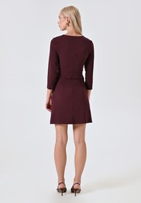 Abito borgogna con maniche a tre quarti, vita aderente e zip sul retro. Il tessuto sembra liscio con una sottile consistenza.