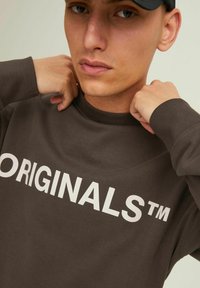 Brown sweatshirt featuring white "ORIGINALS™" text. Made of soft fabric with a crew neckline and ribbed cuffs.