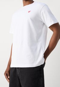 Weißes T-Shirt mit kurzen Ärmeln aus Baumwolle, das ein kleines rotes Grafik-Logo auf der Brust trägt. Der Stoff hat eine glatte Textur.