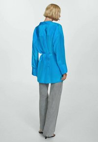 Satijnen blauwe shirt met een strikdetail op de rug, lange mouwen en een rechte snit, gecombineerd met grijze wijde jeans en eenvoudige zwarte sandalen.