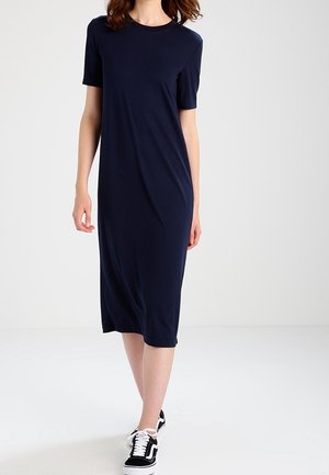 Mujer vestida con un vestido midi azul marino liso de manga corta y zapatillas negras y blancas, de pie sobre un suelo blanco frente a un fondo blanco.