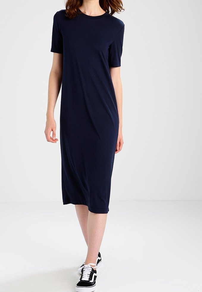 Femme portant une robe midi unie bleu marine à manches courtes et des baskets noires et blanches, debout sur un sol blanc devant un fond blanc.