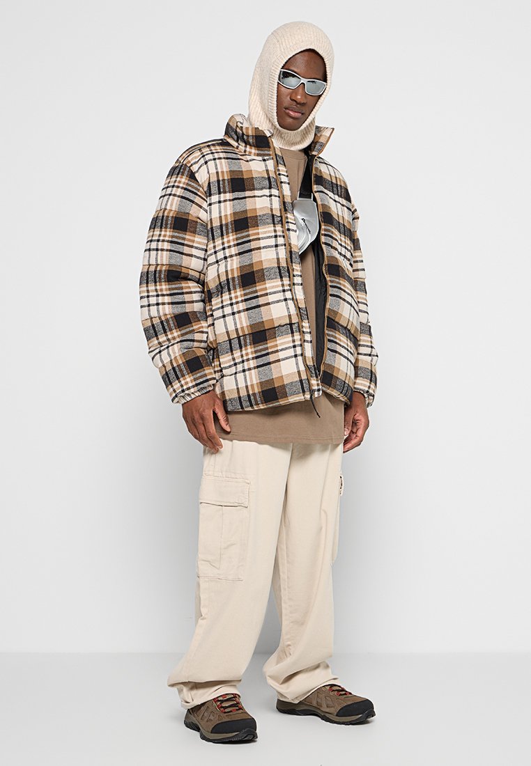 Brązowa i beżowa kurtka puffer w kratę z wysokim kołnierzem, zestawiona z beżowym hoodie i kremowymi spodniami cargo. Szare i czarne buty dopełniają całość.