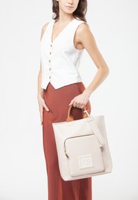 Donna che indossa un gilet bianco senza maniche con bottoni e una gonna color ruggine, mentre tiene una borsa beige con tasca frontale con zip e manici marroni.