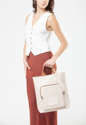 Donna che indossa un gilet bianco senza maniche con bottoni e una gonna color ruggine, mentre tiene una borsa beige con tasca frontale con zip e manici marroni.