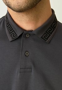 Mørkegrå polo-shirt med en tekstureret sort krave med et geometrisk mønster, to knapper og en quiltet detalje foran.