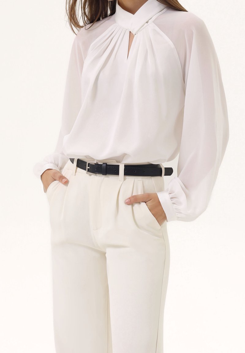 Femme portant un chemisier blanc plissé à manches longues rentré dans un pantalon blanc taille haute avec une ceinture noire, mains dans les poches.