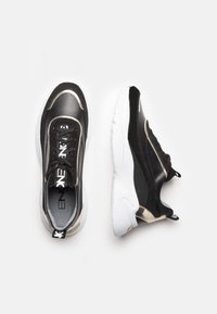 Baskets de sport noires et dorées avec une tige en cuir lisse, semelle en caoutchouc blanche, laçage à l'avant et textures contrastantes sur les côtés.