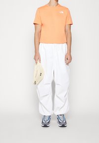 Pomarańczowa krótka koszulka, białe luźne spodnie, szaro-niebieskie sneakersy, żółta nerka z zamkiem błyskawicznym. Model stoi na jednolitym tle.