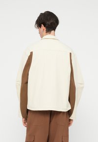 Přehnaná bunda s krémovým hlavním materiálem a hnědými panely na ramenou. Hladká textura, měkký límec a uvolněný střih.