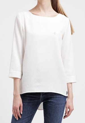 Kobieta ubrana w białą, teksturowaną bluzkę z długim rękawem i dekoltem typu łódka oraz niebieskie dżinsy, stojąca na jednolitym tle.