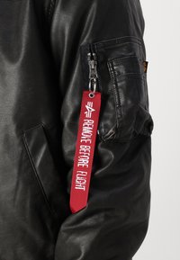 Black leather jacket with a zippered pocket, featuring a red fabric tag with "REMOVE BEFORE FLIGHT" in white text attached to the zipper.