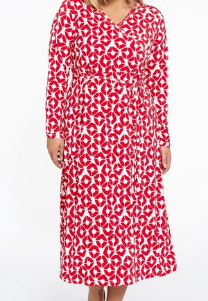 Vrouw draagt een lange mouw, rood-wit patroon wikkeljurk met een gestrikte tailleriem, staand tegen een effen witte achtergrond.