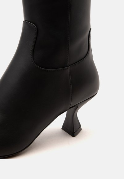 Botte noire en cuir mi-mollet avec talon bloc incurvé et détails cousus sur fond uni clair.