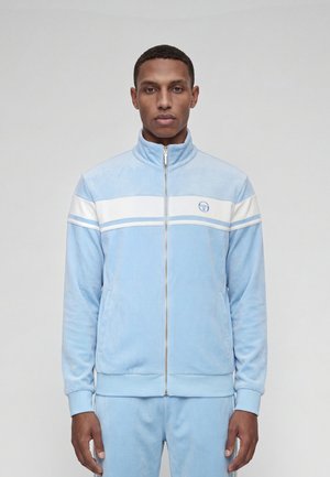 Jeune homme portant une veste zippée bleu clair avec des rayures blanches horizontales sur la poitrine et un pantalon assorti bleu clair.