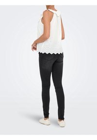 Femme portant un haut blanc sans manches en broderie anglaise avec un ourlet festonné, un jean skinny noir et des chaussures blanches à enfiler, debout sur un fond uni.