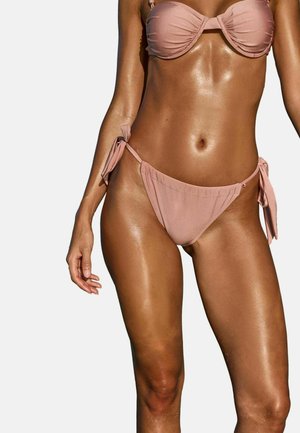 Femme portant un bikini rose clair avec une culotte à liens sur les côtés, montrant le ventre et le haut des jambes sur un fond blanc.