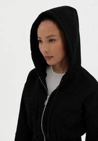 Mustat hupparit, joissa on vetoketju edessä, ylisuuri huppu ja elastinen vyötärö. Pehmeä kangas, jossa on vähäisiäommeltuja yksityiskohtia ja sileä pinta.