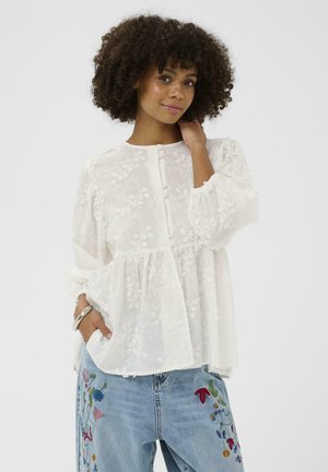 Žena kučeravých vlasov v priesvitnej bielej blúzke s vyšívanými kvetmi a modrých džínsoch s farebnou kvetinovou výšivkou, pózuje s jednou rukou v vrecku.