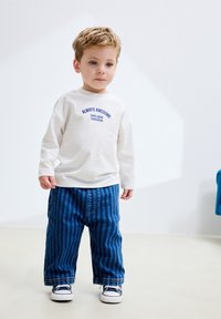 Cămașă albă cu mâneci lungi și text albastru, combinată cu blugi largi cu dungi albastre. Copilul poartă adidași albi. Ținută simplă și modernă.