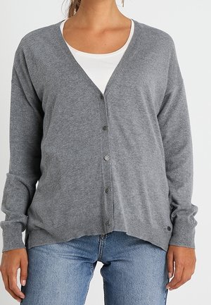 Žena v šedém svetru na knoflíky přes bílý top a světle modré džíny, zobrazena od krku po boky na jednobarevném pozadí.