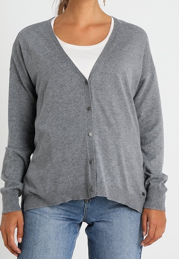 Femme portant un cardigan gris boutonné par-dessus un haut blanc et un jean bleu clair, vue du cou jusqu'aux hanches sur un fond uni.