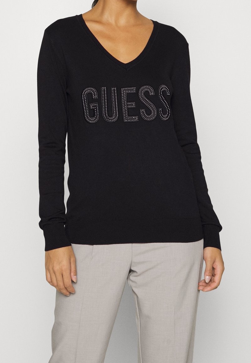 Femme portant un pull à manches longues noir à col en V avec le texte "GUESS" orné, associé à un pantalon gris clair sur un fond uni.