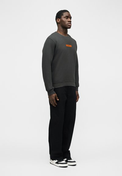 Νέος άνδρας που φοράει σκούρο γκρι φούτερ με πορτοκαλί κείμενο "Voltaire", μαύρο παντελόνι και λευκά-μαύρα αθλητικά παπούτσια, στέκεται μπροστά σε λευκό φόντο.