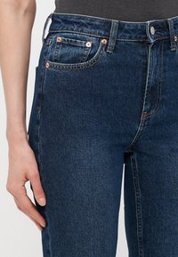 Tmavomodré džínsy z denimu so priliehavým strihom, piatimi vreckami, meďou na hardware a jemnými švami viditeľnými na páse a švoch.