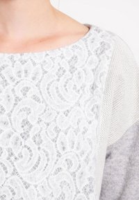 Hellgrauer Pullover mit weißem Spitze-Overlay und kunstvollen, wirbelnden Mustern; texturierte Strickakzente an den Schultern, weiches und lässiges Design.