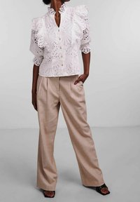 Weiße Bluse mit Lochstickerei, Rüschensäumen und Knopfleiste, kombiniert mit hellbeigen, hochtaillierten Hosen. Flache schwarze Sandalen runden das Outfit ab.