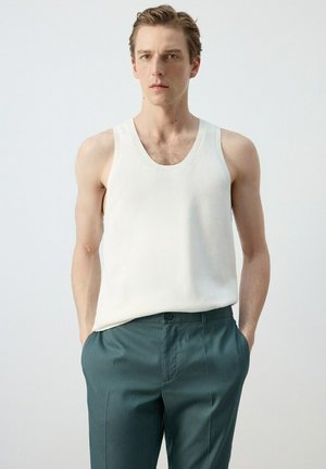 Joven con un top de punto blanco sin mangas y pantalones a medida verde oscuro con las manos en los bolsillos, de pie frente a un fondo liso.