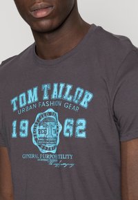 Szara bawełniana koszulka z turkusowym nadrukiem graficznym oraz napisem "Tom Tailor Urban Fashion Gear 1962" i okrągłym logo. Okrągły dekolt.