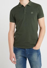 Homme portant un polo à manches courtes vert olive avec le logo "BOSS" sur la poitrine et un jean bleu décoloré avec la main dans la poche.