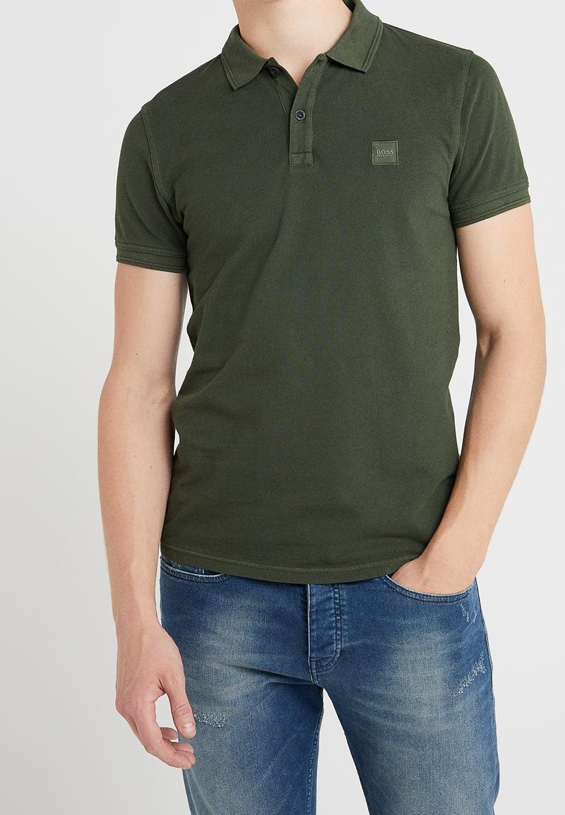 Homme portant un polo à manches courtes vert olive avec le logo "BOSS" sur la poitrine et un jean bleu décoloré avec la main dans la poche.