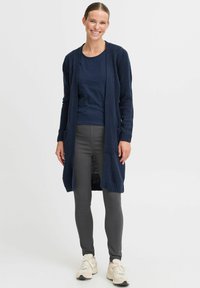 Mornaric plesti cardigan z odprtim sprednjim delom, rebrastimi manšetami in žepi, nosi se preko temnega topa in sivih legic, dopolnjen s bež supergami.