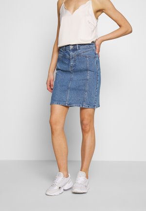 Mujer con blusa blanca sin mangas, falda vaquera azul con costuras frontales y zapatillas blancas, de pie con una mano en la cadera.