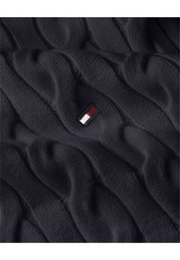 Tissu tricoté bleu marine avec un motif en torsade, présentant un petit emblème avec des rayures rouges, blanches et bleues près du bas. Texture douce.