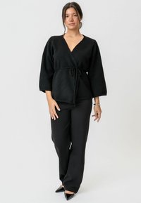 Cardigan negru cu un material tricotat texturat, cu mâneci largi și talie legată, asociat cu pantaloni largi negri și pantofi cu vârf alungit.