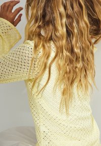 Hellgelber, gestrickter Pullover mit lockerer Passform und strukturiertem Muster. Langes, welliges Haar, das teilweise über eine Schulter sichtbar ist.
