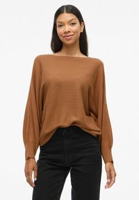 Haut marron à manches longues côtelé avec un décolleté large, coupe décontractée et tissu texturé. Associé à un jean noir taille haute.