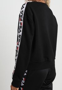 Černá mikina s kapucí, s bílým a červeným značkovým páskem podél rukávů, žebrovanými manžetami a spodním lemem, a volným střihem. Hladká textilie.